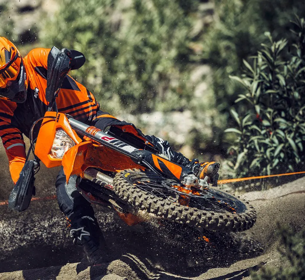 A rider in orange gear sliding an orange dirt bike through a dusty corner